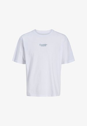 Jeune personne debout et souriante, portant un t-shirt blanc à motif graphique, un short noir à fines rayures, des chaussettes blanches de marque et des baskets blanches.