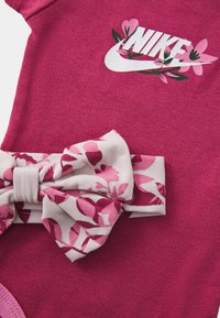 Bodysuit de algodão rosa com um logotipo da Nike branco decorado com elementos florais, complementado por um laço com padrão de flores na cintura.