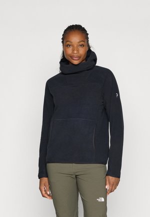 Naine, kes kannab musta kõrge kaelusega fliisjopet ja oliivrohelisi pükse, millel on The North Face'i logo, seisab ühevärvilise tausta ees.