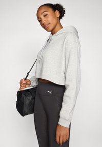 Felpa grigia corta con cappuccio e lacci bianchi, abbinata a leggins neri a vita alta con un piccolo logo. Porta una borsa nera.