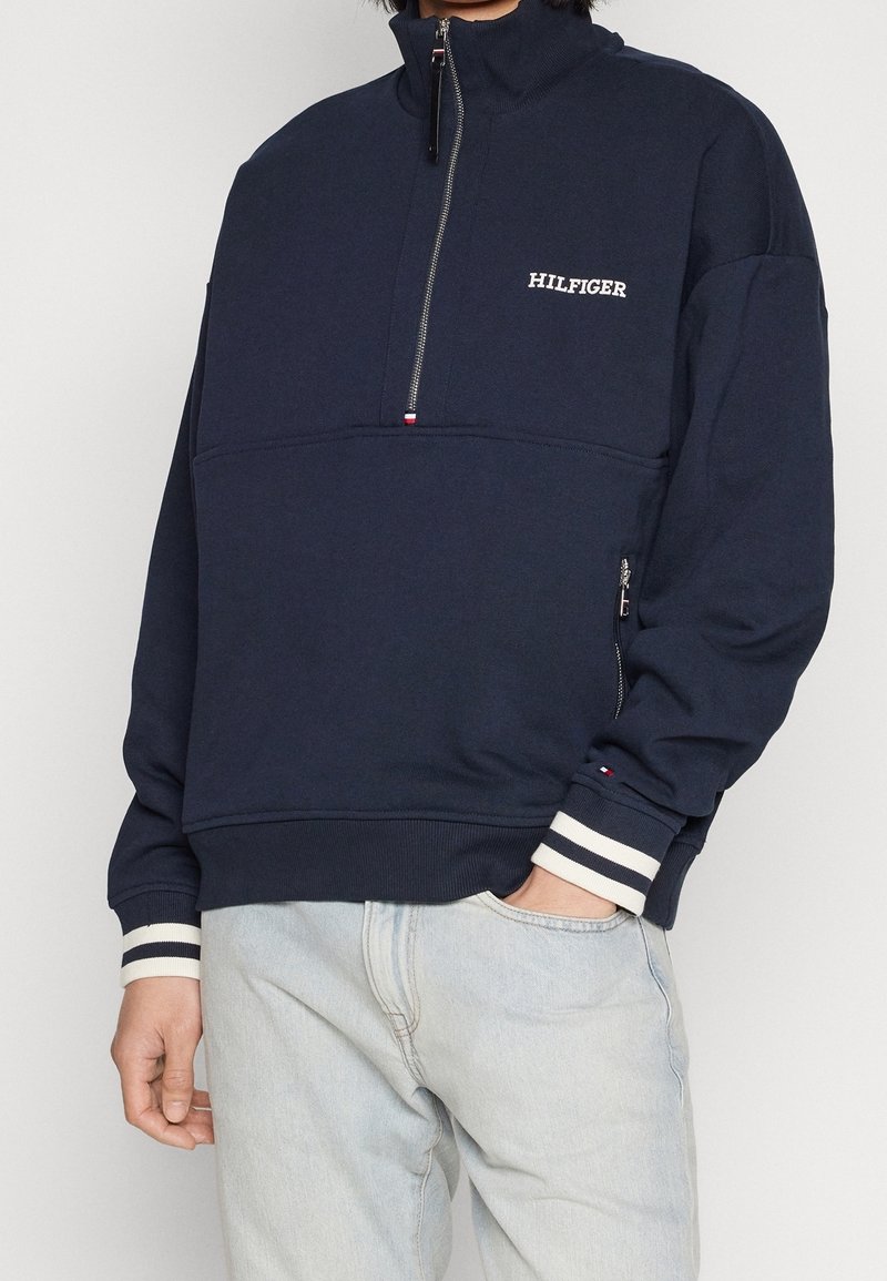 Personne portant un sweat-shirt bleu marine à fermeture éclair partielle avec le texte "HILFIGER", des poignets rayés et un jean bleu clair avec une main dans la poche.