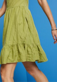 Femme marchant dans une robe sans manches vert clair texturée avec un ourlet à volants et un bracelet argenté au poignet droit, sur un fond bleu.