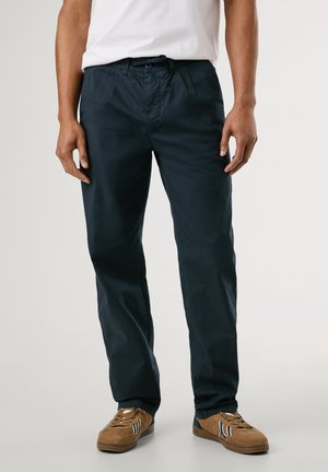 Mand iført sorte afslappede bukser, hvid t-shirt og brune snørebåndssneakers, stående mod en ensfarvet lys baggrund.