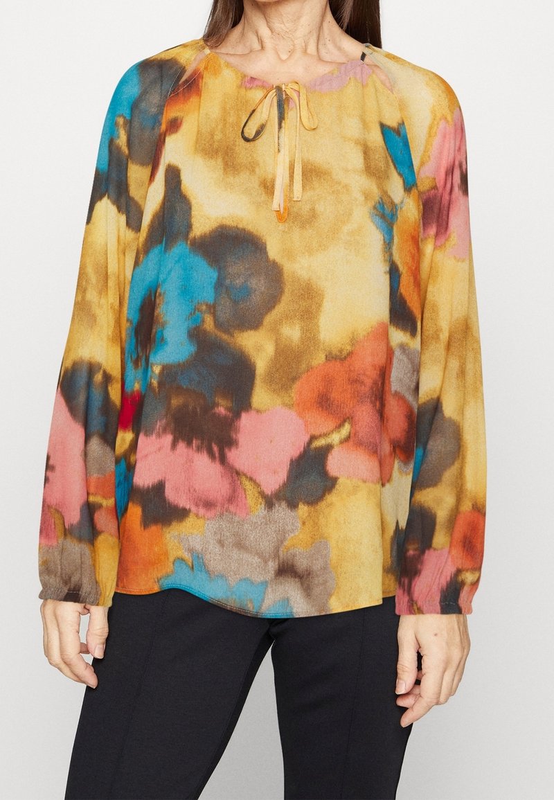 Blouse fluide à manches longues avec un motif floral multicolore dans des teintes de jaune, bleu, rose et marron ; présente un col rond et un détail à nouer.