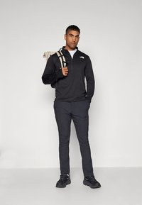 Egy férfi áll szürke háttér előtt, fekete North Face pulóvert, sötét nadrágot és fekete sportcipőt visel, egyik vállán bézs színű hátizsákot hord.