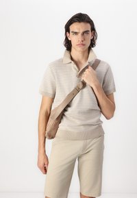 Beige strikket polo skjorte med krage, kombinert med lysebeige shortser. Modellen holder en lysbrun crossover-veske med en glatt tekstur.