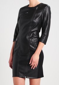 Schwarzes Lederkleid mit drei Viertelarmen, asymmetrischen Reißverschlussdetails und zwei seitlichen Reißverschlusstaschen; figurbetonter Schnitt, glatte Textur.