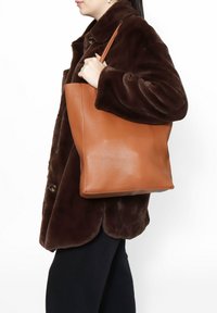Borsa a tracolla in pelle tan con una texture liscia, forma rettangolare e una lunga tracolla, tenuta da una persona che indossa un cappotto di pelliccia marrone.