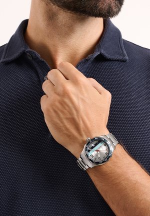 Mann richtet Kragen und trägt eine silberne Metallarmbanduhr mit blauem und weißem Zifferblatt, schwarzen Ziffern und rotem Sekundenzeiger am linken Handgelenk.