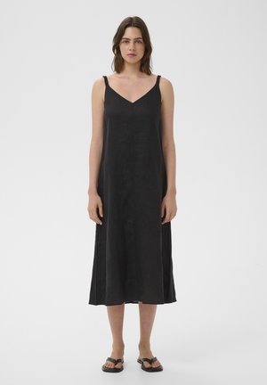 Femme debout portant une robe midi noire sans manches à fines bretelles, associée à des sandales plates noires, sur un fond blanc uni.