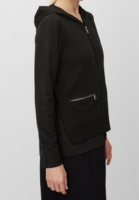 Black zip-up hoodie with a textured fabric, featuring a hood, two zippered pockets, and long sleeves, shown from the side against a neutral background.