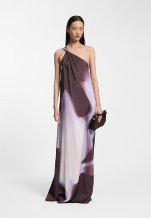 Kobieta w brązowo-białej gradientowej sukni na jedno ramię trzymająca małą czarną kopertówkę na jednolitym tle.