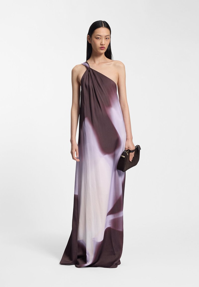 Γυναίκα με φόρεμα καφέ και άσπρης διαβάθμισης με έναν ώμο, κρατώντας μικρό μαύρο τσαντάκι, σε απλό φόντο.