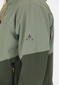 Veste légère verte avec un design bicolore, dotée d'une poche zippée et d'un logo gris foncé sur la manche. Fabriquée à partir d'un matériau lisse et respirant.