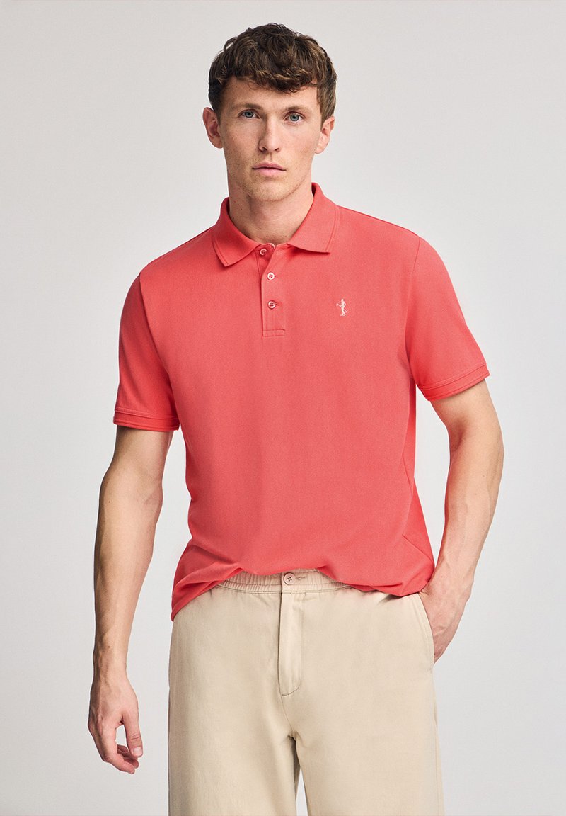 Camisa polo de manga corta color coral con collar y tres botones, que presenta un pequeño logo blanco en el pecho. Combinada con pantalones beige claros.