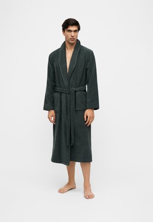 Homme debout pieds nus portant un peignoir gris foncé avec poches et ceinture nouée sur un fond clair uni.