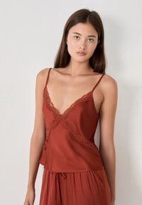 Modelo con cabello largo y lacio que lleva una camiseta de tirantes color óxido con encaje, mostrando un fondo neutro. Iluminación natural suave.