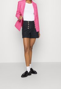 Růžový tkaný blazer se zlatými knoflíky, kombinovaný s tmavými šortkami s vysokým pasem, které mají strukturovaný vzor a další akcenty se zlatými knoflíky.