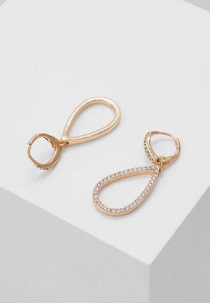 Paire de boucles d'oreilles créoles en forme de goutte d'or, l'une simple et l'autre incrustée de petites pierres claires, exposées sur une surface blanche.