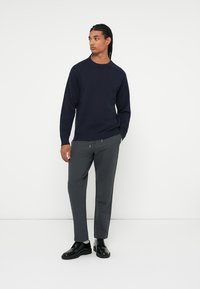 Pull en maille bleu marine avec un col rond, associé à un pantalon gris ajusté. Les chaussures sont noires avec une finition lisse, sur un fond blanc uni.