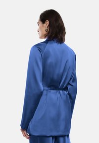Blazer di raso blu con vita cintata, maniche lunghe e finitura liscia. Presenta una silhouette sartoriale e pieghe sottili sul retro.