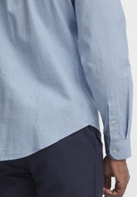 Light blue long-sleeve button-up shirt with a textured fabric and pointed cuffs. Features a curved hem and is paired with dark navy pants.