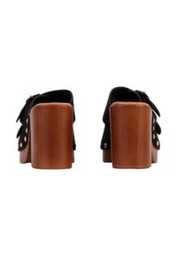 Mules à talons en daim noir avec talons blocs en bois, clous dorés le long des bords supérieurs et sangles à boucle, vus de dos sur fond blanc.