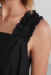 Svart ärmlös topp med volangdetaljer på axelbandet, tillverkad av slät tyg med matt finish. Rena linjer och en fittad passform.