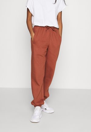 Femme portant un pantalon de jogging couleur rouille avec cordon de serrage et chevilles resserrées, des baskets blanches et un haut blanc ample à manches courtes, debout les mains dans les poches.