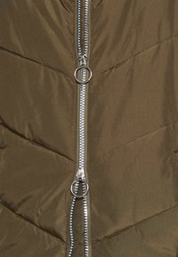 Doudoune vert olive avec une double fermeture éclair argentée à tirette en anneau, texture matelassée et design moderne et épuré.