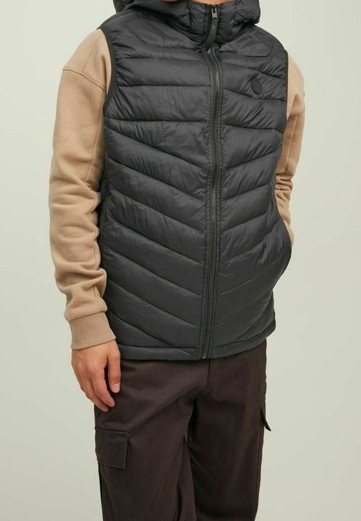 Black insulated vest with a hood, chevron pattern, front zipper, and logo. Paired with a beige long-sleeve shirt and dark pants.