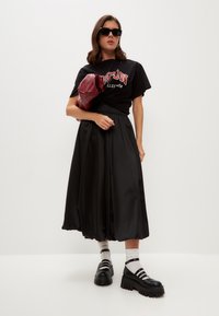T-shirt negru oversized cu text grafic, fustă midi din satin negru, pantofi cu talpă groasă negri și o geantă roșie. Ochelarii de soare completează look-ul.