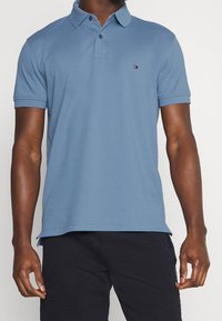 Ljusblå pikétröja i bomull med krage, korta ärmar och en liten logotyp på vänster bröst. Slät textur.