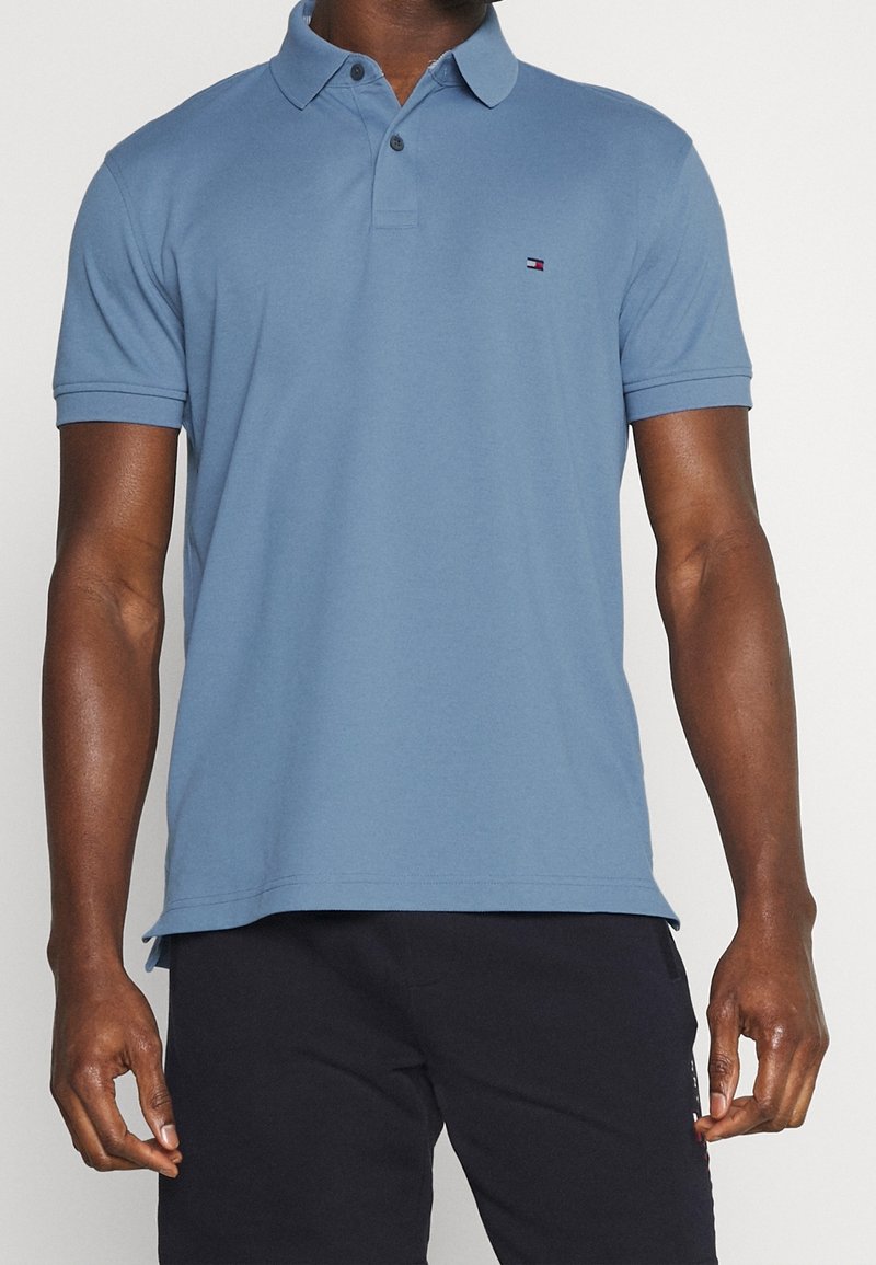 Ljusblå pikétröja i bomull med krage, korta ärmar och en liten logotyp på vänster bröst. Slät textur.