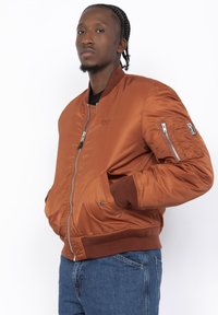 Bronzen bomberjack met een gladde, glanzende textuur. Beschikt over een ritssluiting aan de voorkant, ribgebreide manchetten en een ritszak op de mouw. Gekoombineerd met een blauwe jeans.