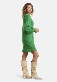 Vestito maglione verde a trecce con maniche lunghe e spacchi laterali, abbinato a stivali beige al ginocchio, che mette in risalto una vestibilità rilassata e un materiale testurizzato.