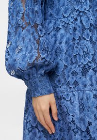 Κυανή δαντελένια φόρεμα με λουλουδάτα μοτίβα, υφή, φουσκωτά μανίκια και εφαρμοστό μπούστο. Περιλαμβάνει πολύπλοκες λεπτομέρειες από δαντέλα.