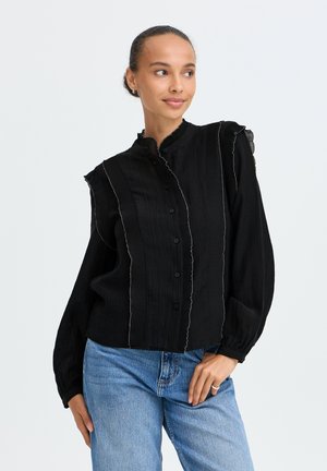 Femme aux cheveux attachés portant un chemisier noir boutonné avec des volants plissés, associé à un jean bleu clair, regardant légèrement sur le côté.