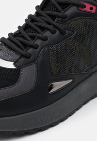 Chaussure de sport noire composée d'un mélange de matériaux, incluant du cuir lisse et du mesh. Des accents texturés et des lacets noirs complètent le design.