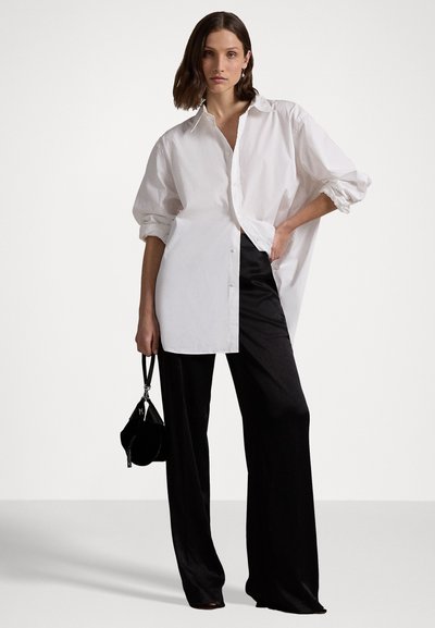 Υπερμεγέθη λευκή πουκάμισο με αναδιπλωμένα μανίκια, συνδυασμένο με ρέοντες μαύρους παντελόνες και μια μικρή μαύρη τσάντα, που αναδεικνύει έναν απλό σχεδιασμό.