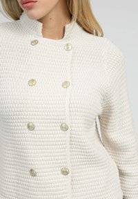 Cardigan blanc texturé avec un col montant, présentant un design à double boutonnage et des boutons dorés.