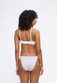 Γυναίκα με σγουρά μαλλιά που φοράει λευκό σουτιέν και ταίριαζα εσώρουχα, εμφανιζόμενη από πίσω σε λευκό απλό φόντο.