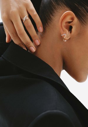 Oreja derecha con pendientes modernos de plata con diseño en zigzag y colgantes, mano con anillos de oro tocando el cuello, vistiendo un blazer negro.