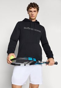 Un jeune homme portant un hoodie noir Björn Borg et un short blanc tient une raquette de tennis et une balle de tennis jaune devant un fond uni.