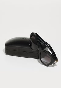 Lunettes de soleil noires en forme d'œil de chat, avec une monture en plastique lisse, des verres foncés et une finition dorée sur les tempes, reposant sur un étui noir.