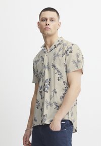 Camisa de botones de manga corta en color beige con estampados de flores azules y mariposas, con un corte relajado y un diseño de cuello con muesca.
