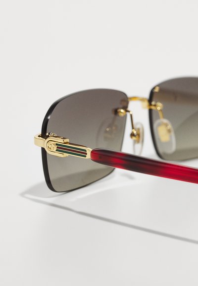 Gafas de sol con un marco cuadrado negro, lentes grises, acentos metálicos dorados y una patilla a rayas rojas y verdes. Las patillas son de plástico rojo.