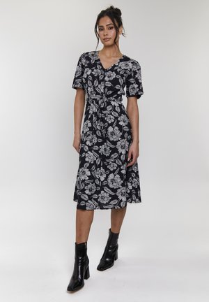 Robe noire à motifs floraux avec un décolleté en V, des manches courtes et une taille cintrée. Portée avec des bottines noires à chevilles. Tissu lisse et longueur au genou.