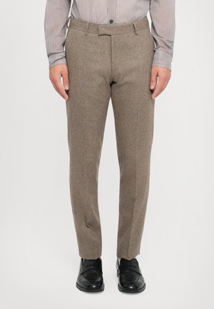 Homme portant un pantalon marron sur mesure, une chemise rayée boutonnée et des chaussures habillées en cuir noir, debout sur fond blanc.