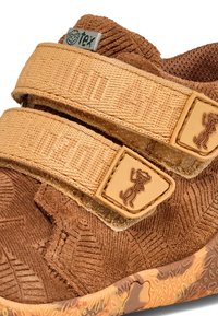 Zapato de tela marrón con correas de Velcro. Parte superior texturizada, suela con patrón y un pequeño logo de mono en las correas. Diseño ligero.